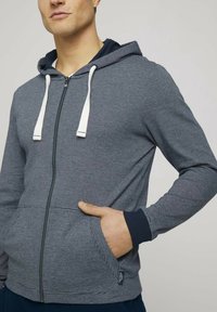 Felpa grigia con zip, presentante cordini bianchi, texture a righe, polsini a coste e tasche laterali. Contrasto di colore notevole con fodera interna blu navy.