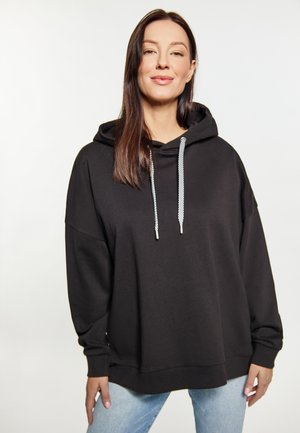 Kvinde iført en løs sort hættetrøje med hvide snore og lyseblå jeans, stående mod en ensfarvet hvid baggrund.