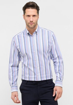 Mand iført en blå, hvid og rød skjorte med lodrette striber og mørke bukser, stående foran en ensartet lys baggrund.