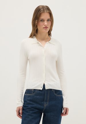 Donna che indossa un cardigan bianco a coste con bottoni e jeans blu scuro, in piedi davanti a uno sfondo chiaro e uniforme.