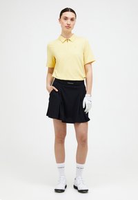Polo shirt giallo a maniche corte con dettaglio zip, abbinato a una gonna plissettata nera. Indossa un guanto da golf bianco e scarpe sportive.