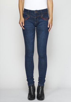 Žena v tmavě modrých úzkých džínách s červenou květinovou výšivkou u kapes a černých kotníkových botách, stojící před jednobarevným pozadím.