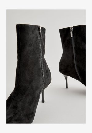 Fekete szarvasbőr bokacsizma elegáns kialakítással, oldalsó cipzárral és magas tűsarkúval, sima textúrával és modern sziluettel.
