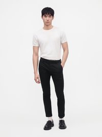 Homme debout portant un t-shirt blanc à manches courtes, un pantalon noir slim, des chaussettes blanches et des mocassins noirs sur un fond blanc uni.