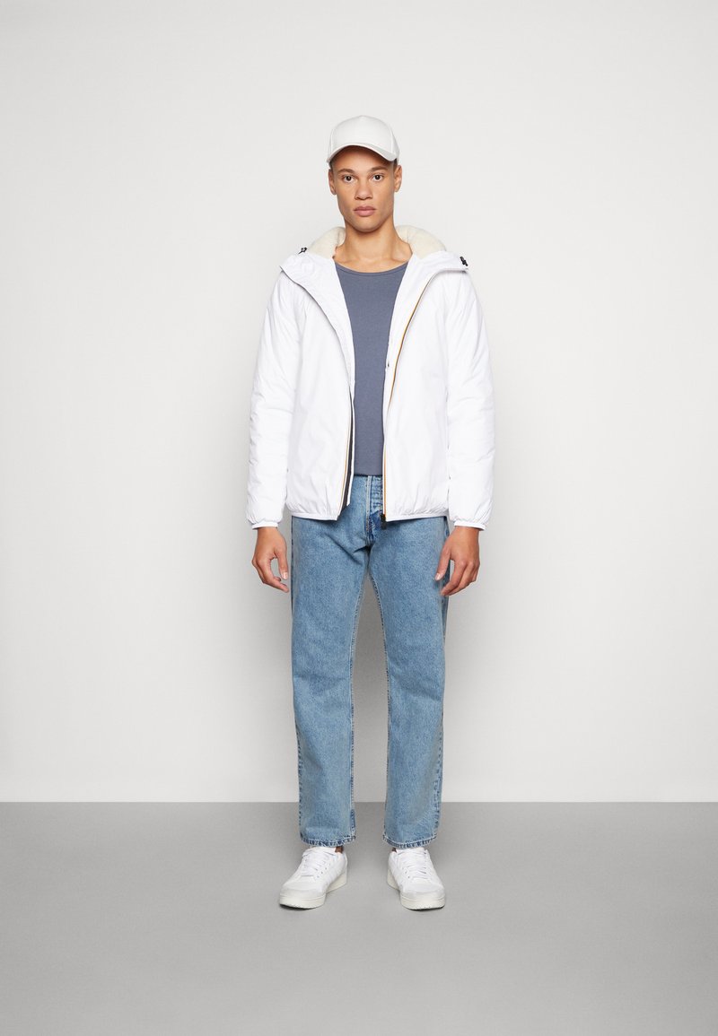 Giacca puffer bianca con foderatura beige, abbinata a una maglietta navy e jeans azzurri chiari. Il modello indossa un cappellino bianco e sneakers bianche.