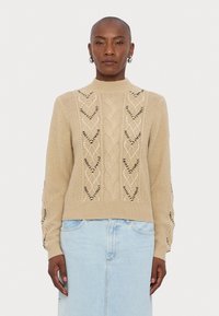 Beige kudumüts, millel on kõrge kaelus, kabeli kudumise mustrid ja mustrilised aktsendid. Kombineeritud helesinise teksaseelikuga.