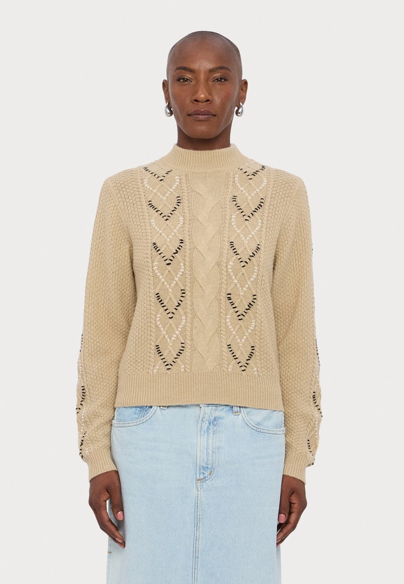 Beige kudumüts, millel on kõrge kaelus, kabeli kudumise mustrid ja mustrilised aktsendid. Kombineeritud helesinise teksaseelikuga.