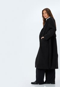 Czarny wełniany długi płaszcz z paskiem w talii, szerokimi rękawami i kieszeniami bocznymi, noszony na luźnych czarnych spodniach. Gładka tekstura, minimalistyczny design.