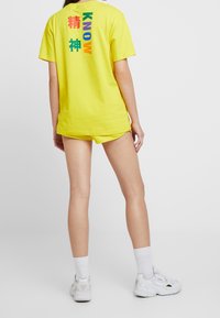 Gul bomullströja med färgglad text på ryggen, matchande shorts och vita sneakers. Enkelt mönster, avslappnad passform.