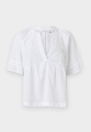 Λευκό κοντομάνικο πουκάμισο με V-λαιμόκοψη, κεντημένα λουλούδια στο στήθος και βολάν στις μανσέτες, κατασκευασμένο από ελαφρύ ύφασμα.