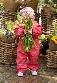 Rød vandtæt heldragt med elastiske håndled, holder grønne krydderurter og pink blomster, står ved kurve af flettet rattan fyldt med farverige blomster.