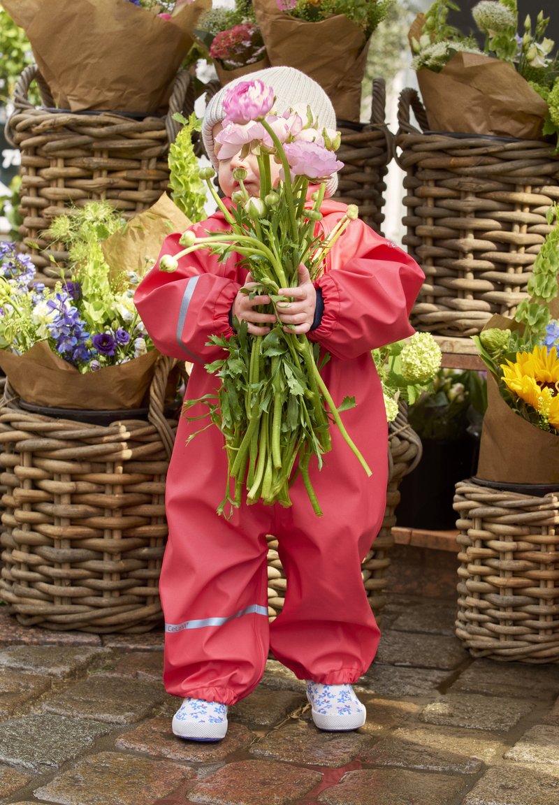 Rød vandtæt heldragt med elastiske håndled, holder grønne krydderurter og pink blomster, står ved kurve af flettet rattan fyldt med farverige blomster.