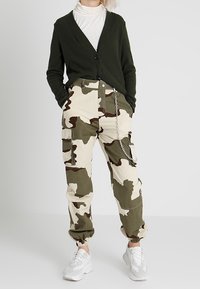 Grüner gerippter Cardigan mit V-Ausschnitt und schwarzen Knöpfen, kombiniert mit beigen camouflage Cargo-Hosen mit Taschen und elastischen Bündchen. Weiße Sneakers.