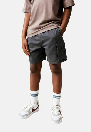 Graue Cargo-Shorts mit Seitentaschen, kombiniert mit weißen Sneakers mit kastanienbraunen Akzenten und blau gestreiften Knöchelsocken. Schlichtes T-Shirt-Design.