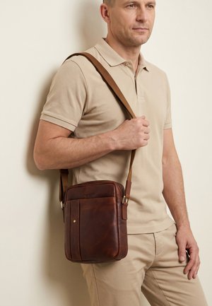 Homme en polo beige et pantalon tenant un sac en cuir marron porté en bandoulière contre un mur clair.