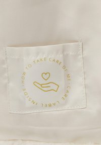 Weißes Pflegeetikett aus Stoff mit goldenen Texten und Grafiken: eine Hand mit einem Herzen, Pflegeanweisungen und einem kreisförmigen Randdesign.