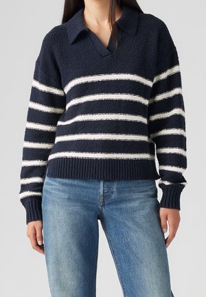Femme portant un pull en maille bleu marine avec des rayures blanches horizontales et un col, associé à un jean bleu clair, debout devant un fond gris.