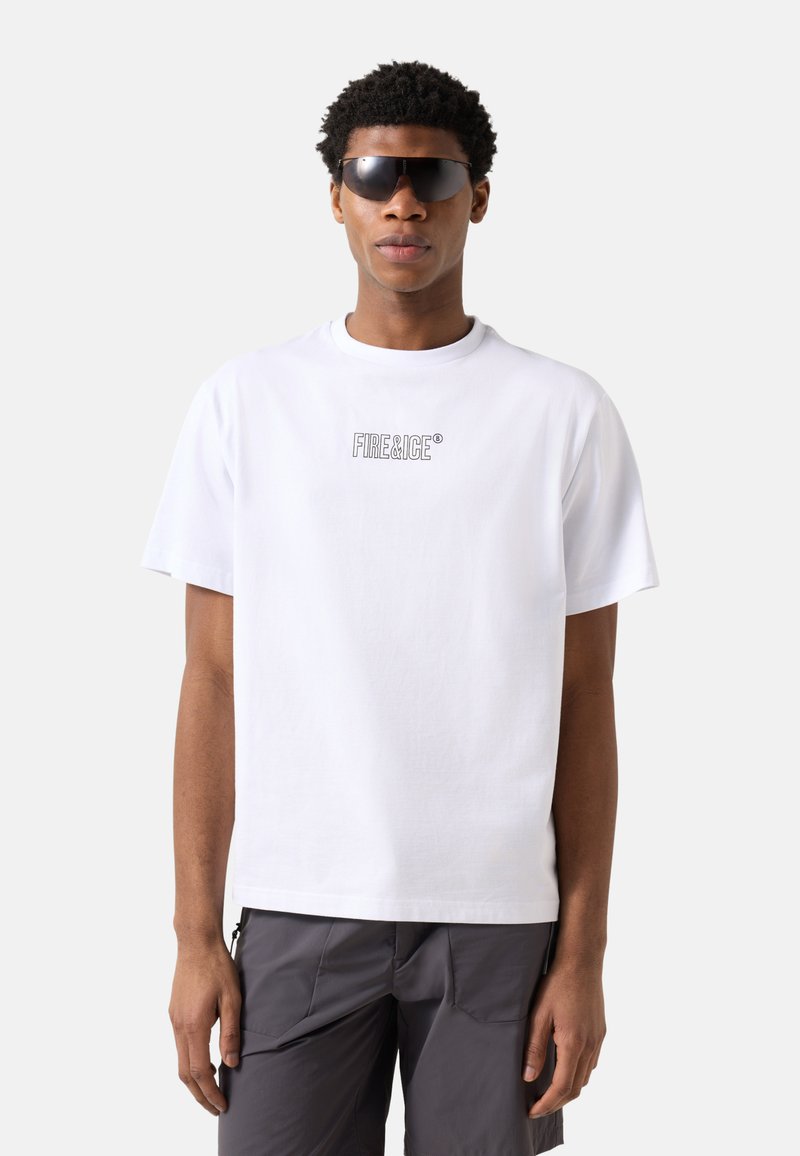 Homme portant des lunettes de soleil noires, un t-shirt blanc "FIRE & ICE" et un pantalon gris foncé, debout devant un fond clair uni.