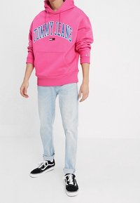 Sudadera rosa con texto "TOMMY JEANS" en azul, bolsillo tipo canguro en la parte frontal, combinada con jeans azul claro y zapatillas negras con suelas blancas.
