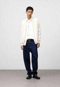 Maglione bianco a trama, camicia bianca con bottoni, jeans larghe blu navy, cintura nera, stivali alla caviglia neri. Sfondo neutro con pavimento grigio.