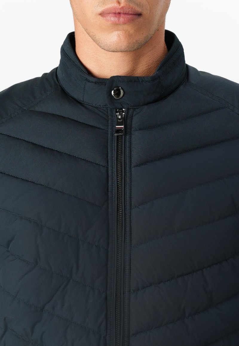 Ciemnoniebieska ocieplana kurtka z wysokim kołnierzem, poziomymi przeszyciami, zamkiem błyskawicznym z przodu i napą przy kołnierzu.