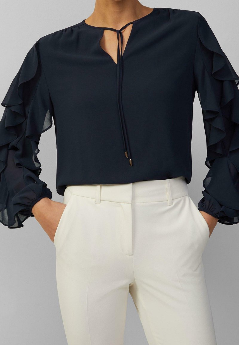Femme portant un chemisier bleu marine avec des manches volantées et un col noué, associé à un pantalon blanc taille haute, mains dans les poches.