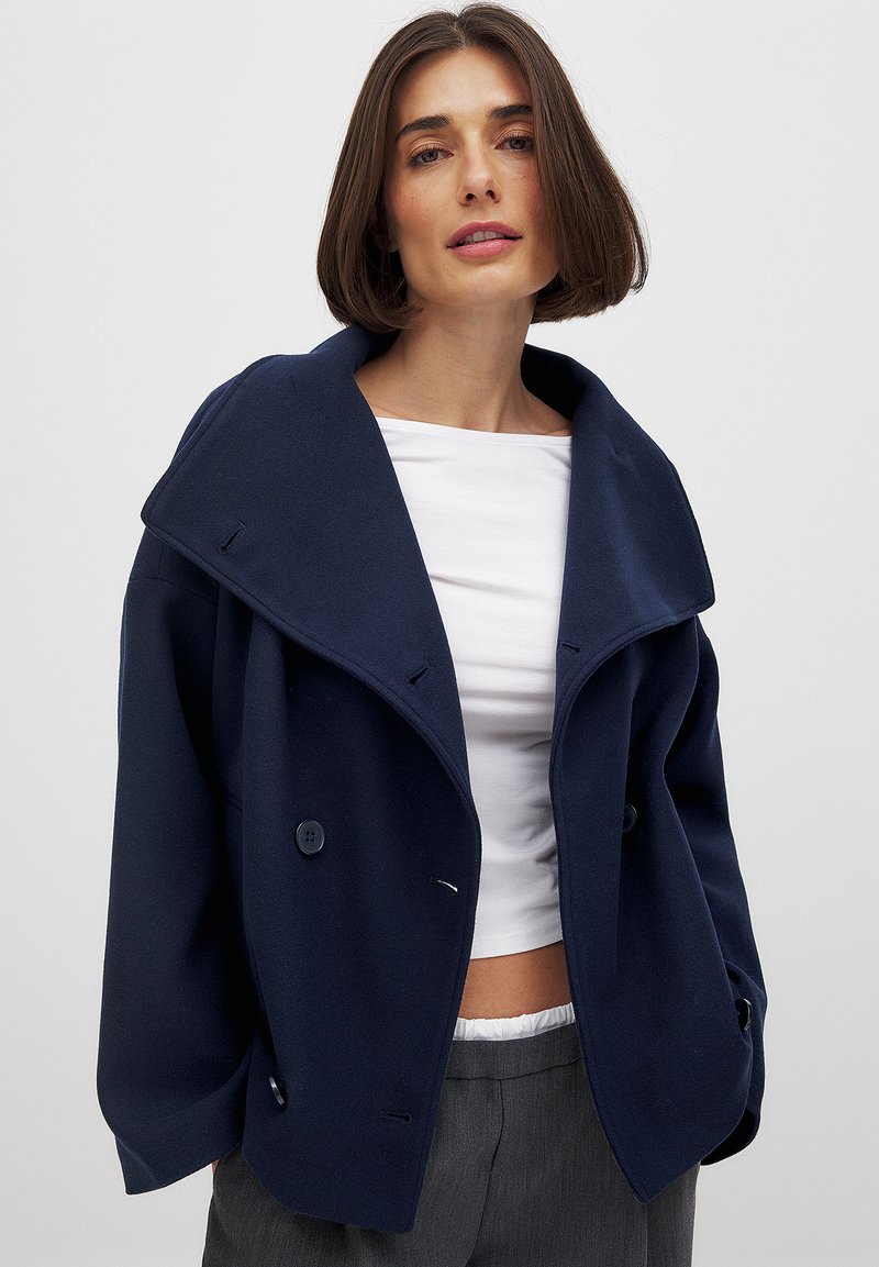 Tumedast sinine kahekordne mantel laia kraega, tekstureeritud kangas, suured nööbid ja laiad varrukad. Kombineeritud valge ülakehaga ja hallide pükstega.