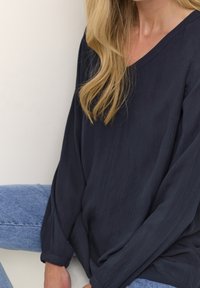 Frau mit langen blonden Haaren, die eine lockere schwarze V-Ausschnitt-Bluse und hellblaue Jeans trägt, sitzt vor einem schlichten Hintergrund.