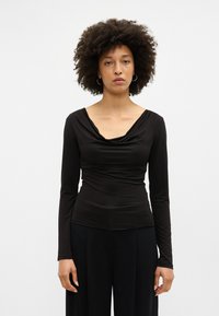 Črna dolga majica z drapirano cowl izrezom in oprijetim telesom, izdelana iz gladkega blaga, nošena z črnimi hlačami z visokim pasom.