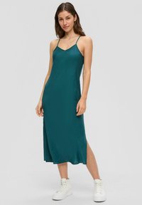 Miętowa satynowa sukienka midi na ramiączkach, z dekoltem w kształcie litery V i rozcięciem po boku, stylizowana z białymi sportowymi trampkami.