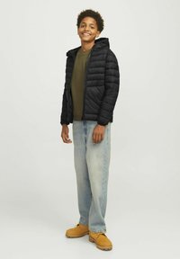Zwarte pufferjack met een capuchon, ritssluiting en geribbelde manchetten, gedragen over een olijfgroen shirt, gecombineerd met lichtblauwe jeans en tan kleurige boots.