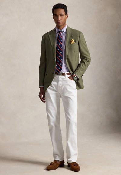 Άνδρας που φοράει χακί πράσινο σακάκι, ριγέ πουκάμισο, ναυτικό γραβάτα με κόκκινες ρίγες, λευκό παντελόνι, καφέ μοκασίνια και κίτρινο μαντηλάκι τσέπης, στέκεται με το ένα χέρι στην τσέπη.