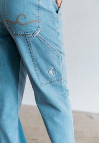 Helle blaue Jeanshose mit lockerem Schnitt, die mit Zickzacknähten, seitlichen Taschen und einem runden bestickten Logo am Bein versehen ist.