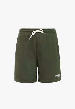 Niño rubio de pie con las manos en los bolsillos, vistiendo una camiseta beige con un logo azul, pantalones cortos verde oscuro, calcetas blancas y zapatillas grises sobre un fondo liso.