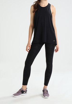 Kobieta ubrana w czarny bezrękawnik sportowy, czarne legginsy z wzorem oraz lawendowe buty do biegania Nike, stojąca na tle jednolitego tła.