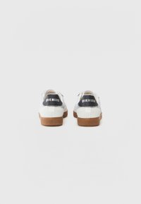 White sneakers with textured fabric, rubber soles, and a brown accent. Includes a black logo on the heel. Classic lace-up design.