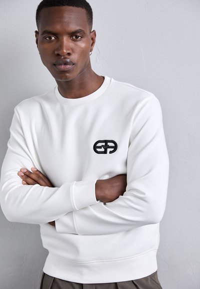 Sweatshirt branca com gola redonda e mangas longas, apresentando um logo bordado preto no peito e punhos canelados. Textura suave.
