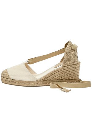Espadrille en toile beige avec une tige en toile, semelle en jute tressé, et lacets pour le soutien de la cheville. Design à dos ouvert et bout rond.