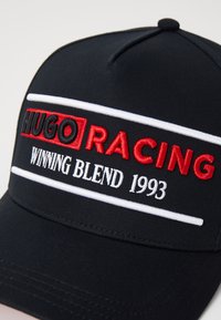 Boné preto de tecido, com o bordado "HUGO RACING" em vermelho, com riscas brancas e "WINNING BLEND 1993" embaixo.