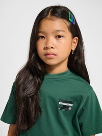 Młoda dziewczyna z długimi ciemnymi włosami, nosząca zielone i niebieskie spinki do włosów, małą kolczyk-kółko i zielony sportowy t-shirt hummel.