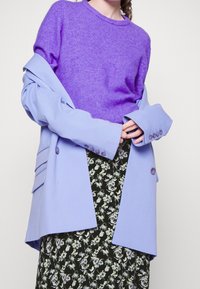Femme portant un pull violet, un blazer oversized bleu clair et une jupe noire à fleurs, se tenant les mains jointes devant elle.