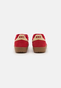 Sarkani zamšādas kedas ar brūnu gumijas zoli. Zelta Nike zīmols uz papēžiem. Zema griezuma dizains ar teksturētām akcentēm. Pastiprināta papēža konstrukcija.