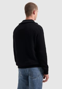 Jonge man met zijn rug naar de camera gekeerd, gekleed in een zwart geribd sweater en lichtblauwe jeans, tegen een effen lichte achtergrond.