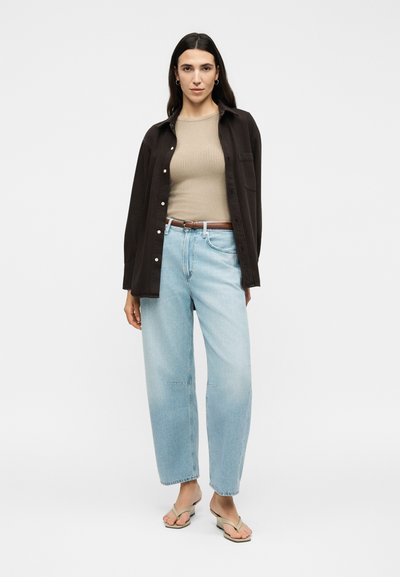 Γυναίκα με μακριά σκούρα μαλλιά που φοράει ανοιχτό μπλε τζιν, μπεζ ριμπ τοπ, σκούρο ανοιχτό πουκάμισο και μπεζ πέδιλα με τακούνι, στέκεται μπροστά σε λευκό φόντο.