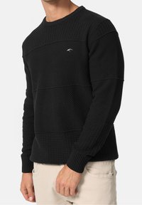 Pull texturé noir avec un col rond, présentant des panneaux horizontaux et des poignets côtelés. Marquage subtil en blanc sur la poitrine.