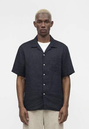 Mann med kort blond hår som har på seg en svart kortermet skjorte med knapper over en hvit t-skjorte og lyse beige bukser, som vender framover mot en ensfarget bakgrunn.