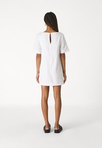 Vestido de lino blanco con mangas cortas, que presenta un pequeño detalle de ojal en la parte de atrás y un corte recto. Llevado con sandalias planas negras.