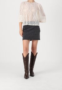 Haut en dentelle blanche à manches 3/4 porté avec une jupe en denim noir ornée de clous. Porté avec des bottes de cow-boy marron.