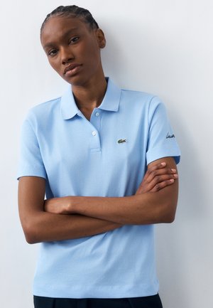 Kvinde med flettet hår, iført en lyseblå polo med en lille grøn krokodille-logo, armene krydset mod en hvid baggrund.