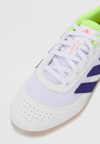 Zapato deportivo blanco con parte superior de malla y acentos sintéticos texturizados. Presenta rayas moradas y un cuello verde brillante. Área del dedo del pie perforada.